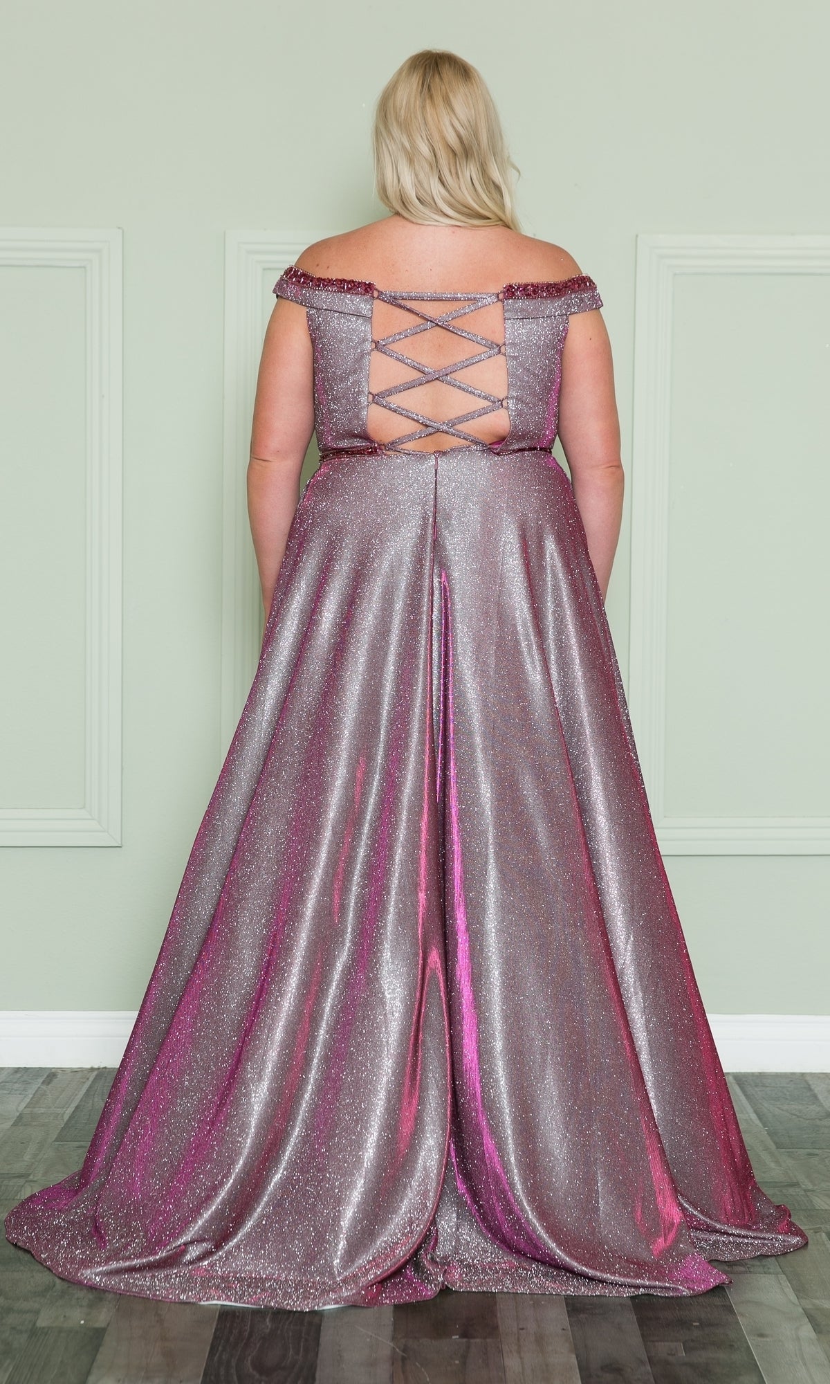 A woman models the Plus-Size Long Glitter-Knit Prom Dress W1096, facing a wall to display the shimmery purple off-the-shoulder gown's lace-up back and sparkling full skirt.